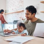A man holding a document works on a laptop with a child sitting on his lap, while a woman is in the background in a kitchen.