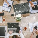 Multiple people seated around a wooden table using laptops, tablets, and phones with charts, papers, and coffee cups.