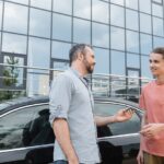A father hands car keys to his teenage son as they stand in front of a building with a black car parked behind them.