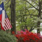 A home's front porch with the American and Texan flag displayed in front of colorful landscaping.