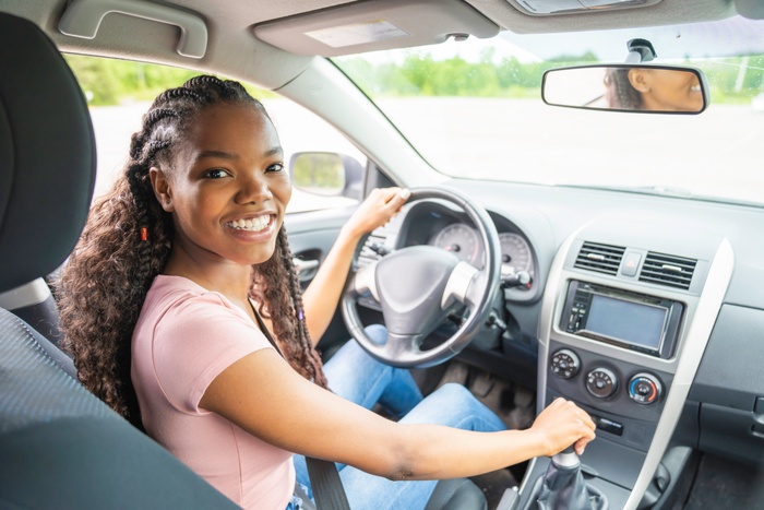 A smiling female teenager sits in the driver's seat with one hand on the wheel and the other on the gear shift.