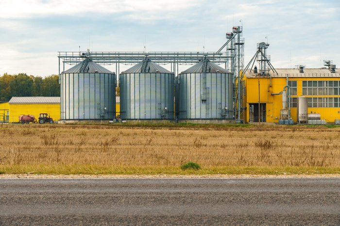 Three grain silos standing beside a bright yellow processing plant across a dry, harvested field under cloudy skies.