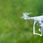 A white quadcopter drone flying outside. The greenery in the background is blurred.
