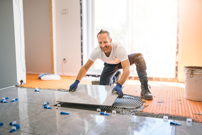 A man kneeling on a tiled floor wearing gloves while carefully placing a large tile onto fresh adhesive.