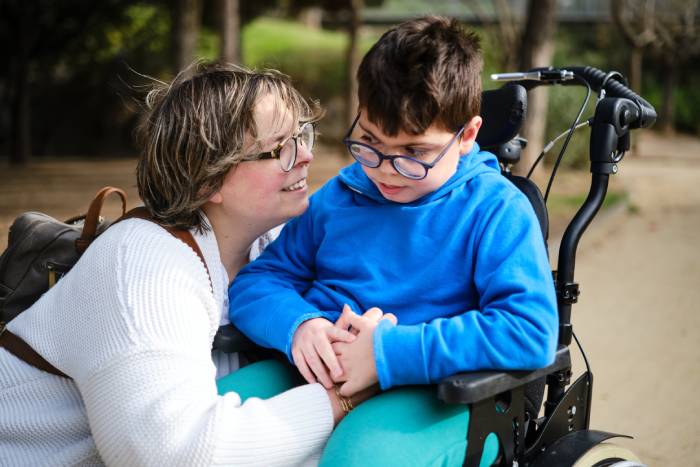 A mother in a white sweater squats down to hug her son, who is wearing a blue sweater and sitting in a black wheelchair.