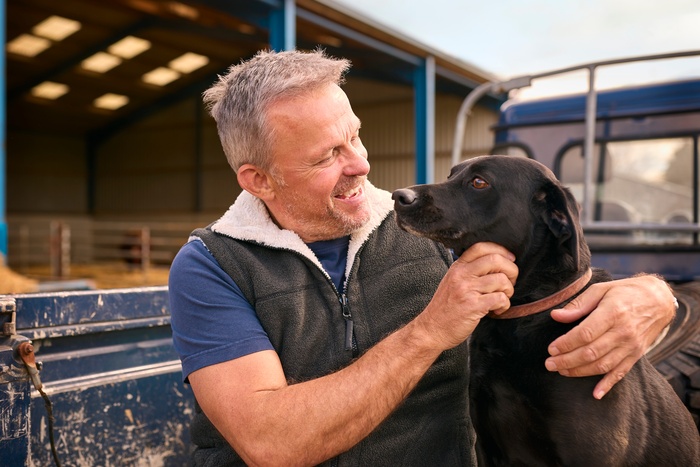 An older man wearing a gray vest smiles while petting his black dog next to a truck in front of a barn.