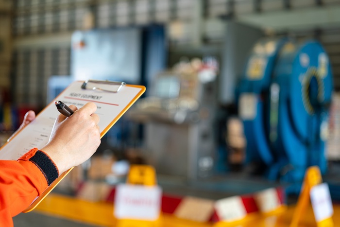 A person wearing an orange safety uniform fills out a checklist on a clipboard inside an industrial warehouse facility.