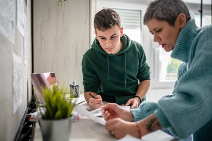 An older person leaning against a counter and teaching an attentive younger person as they look over paperwork together.