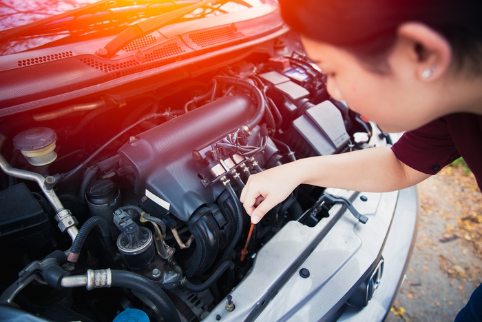 A young woman leans over the open hood of her car and lifts the oil dipstick to check the engine oil.