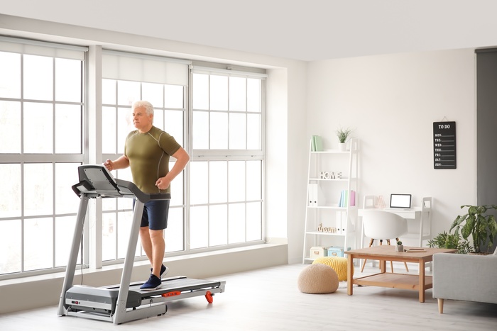 An older man jogging on a treadmill in a living room next to large windows. A desk setup is behind him.