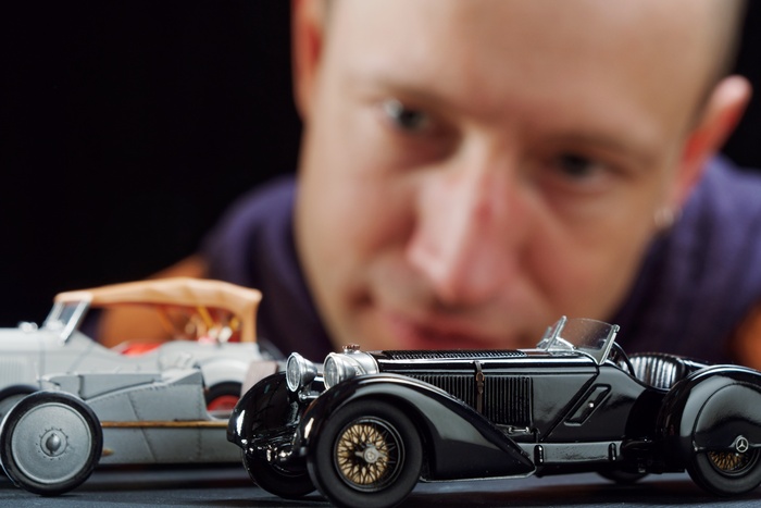 A man leaning in close to examine two vintage diecast model cars, one black and one silver, on a dark tabletop.