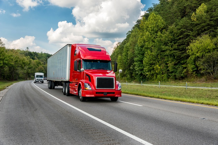 A red semi-truck drives down a highway surrounded by trees. Another car with a trailer trails behind it.