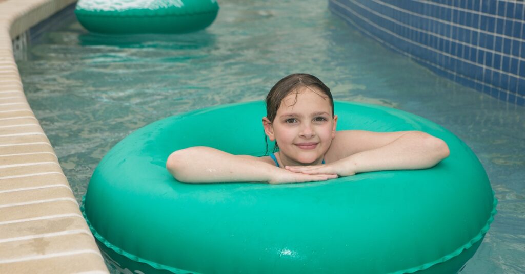 A smiling young girl with wet hair floating in the middle of a circular green inner tube on a lazy river.