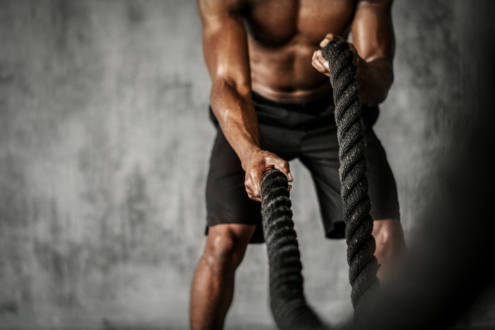 An athletic man wearing black shorts while holding two thick ropes and using them to train for better performance.