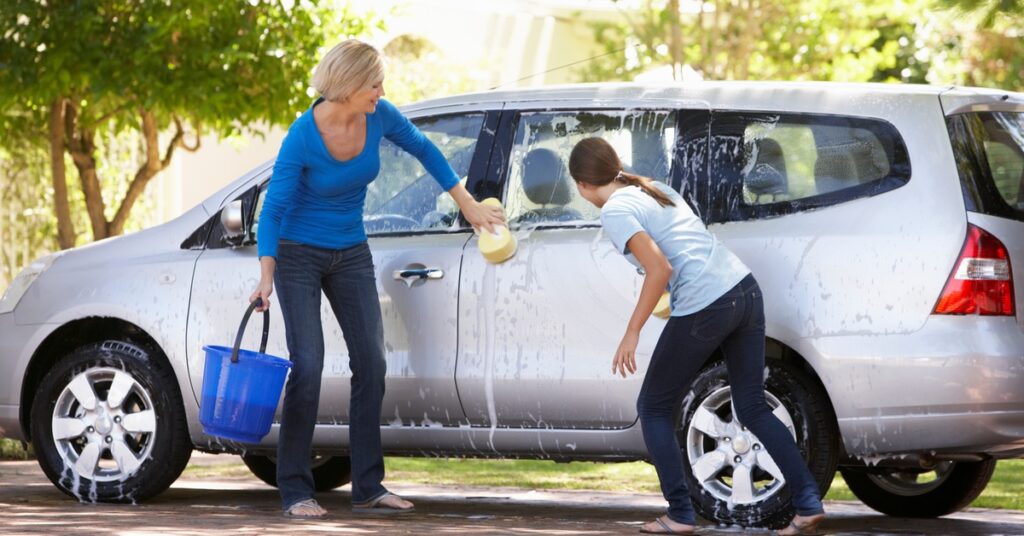 A mother and daughter cleaning a gray car together. Both of them have sponges, and the mom is holding a bucket.