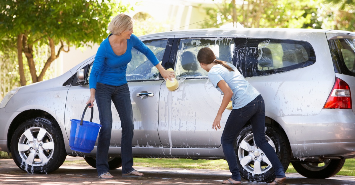 A mother and daughter cleaning a gray car together. Both of them have sponges, and the mom is holding a bucket.
