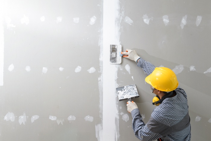 A worker in white gloves and a yellow hard hat using a large taping knife to smooth out plaster on a drywall seam.