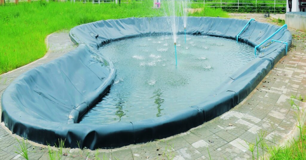 Water splashes from upright blue pipes into a shallow, black-lined outdoor pond surrounded by tile and green grass.