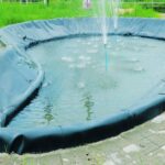 Water splashes from upright blue pipes into a shallow, black-lined outdoor pond surrounded by tile and green grass.