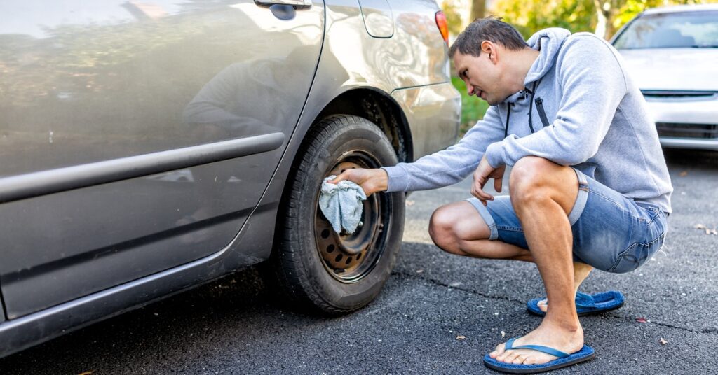 A man in shorts, a hoodie, and flip flops squats down and rubs a cloth against the rusted metal hubcap of his car.