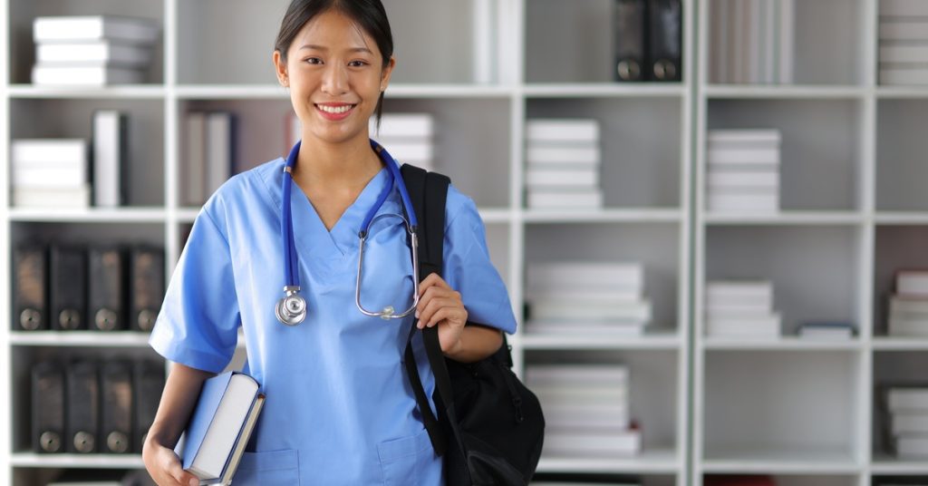 A medical student carries a backpack and holds textbooks. She's also wearing scrubs and a stethoscope around her neck.