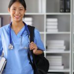 A medical student carries a backpack and holds textbooks. She's also wearing scrubs and a stethoscope around her neck.