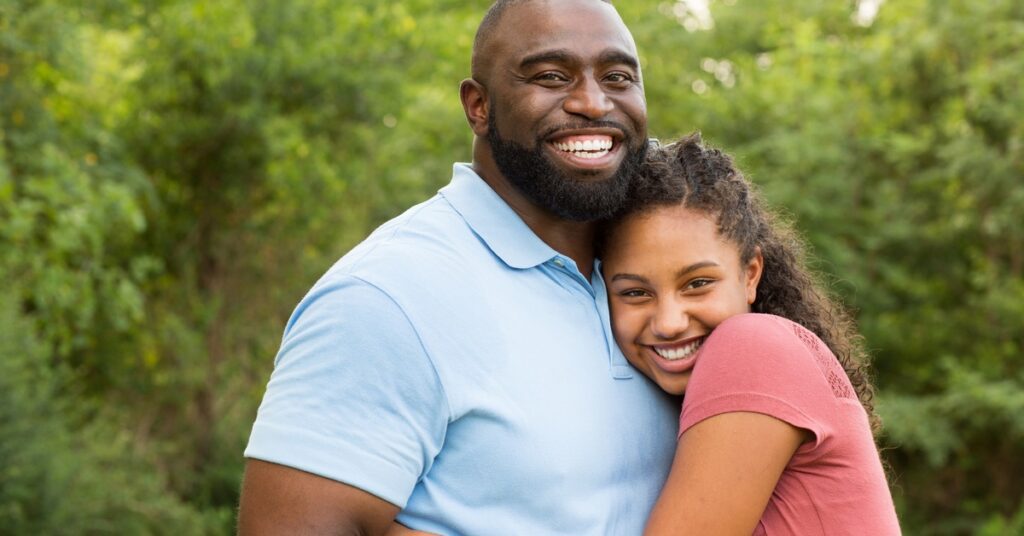 A bearded man in a collared shirt hugging a smiling teenage girl. They're standing in front of trees and bushes.