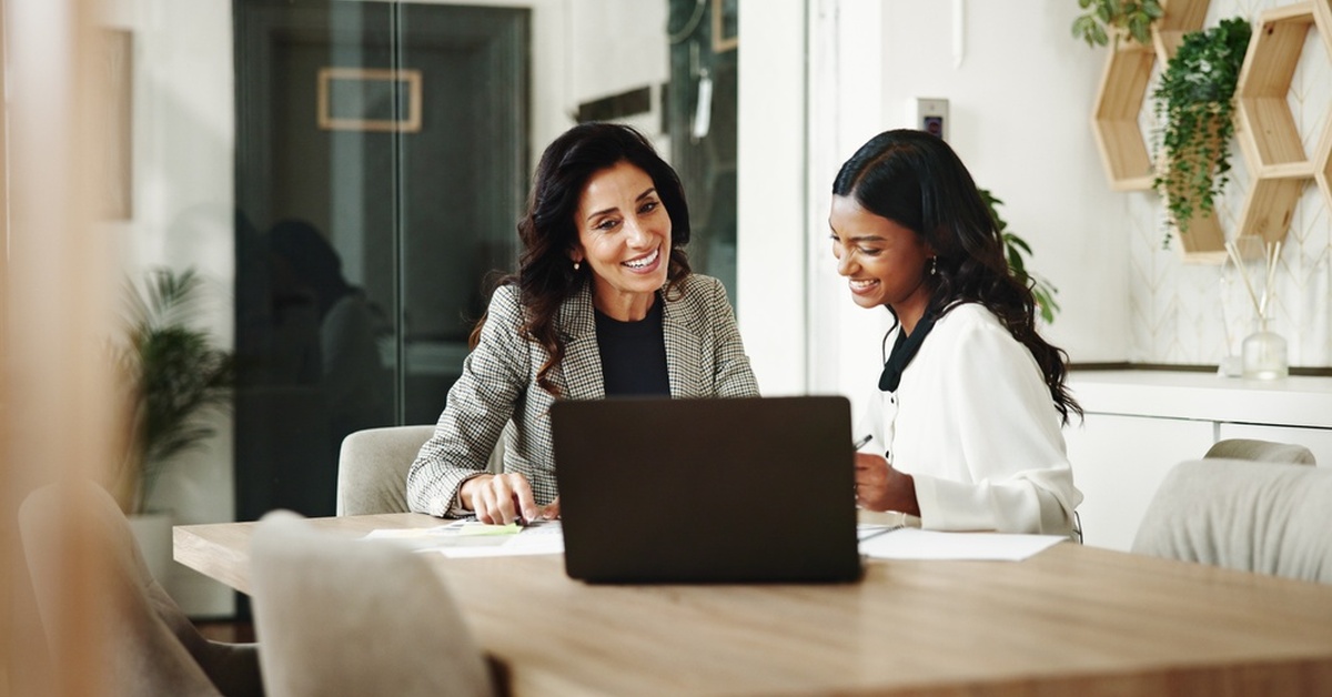 Two professional businesswomen are smiling and using laptops in an office while having a discussion.