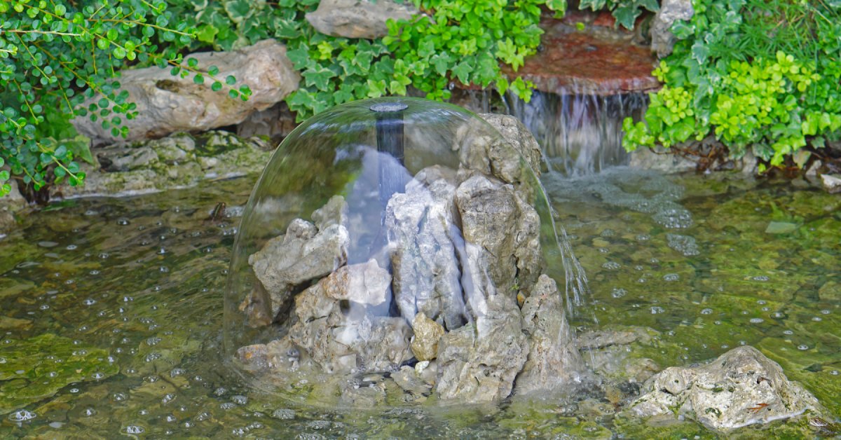 A small pond surrounded by rocks and plants. A fountain in the middle sprays a cascading dome of water over a pile of rocks.