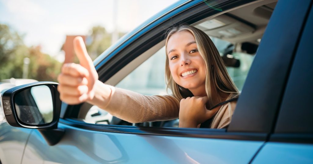 A close-up of a happy young woman, smiling and showing a thumbs-up outside of her car window with her seat belt on.