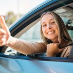 A close-up of a happy young woman, smiling and showing a thumbs-up outside of her car window with her seat belt on.
