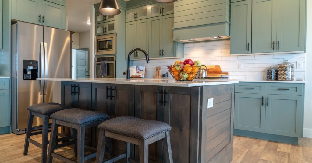 A large L-shaped kitchen features sage-green cabinets and a dark-brown island. Pendant lights hang over the island.