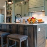 A large L-shaped kitchen features sage-green cabinets and a dark-brown island. Pendant lights hang over the island.