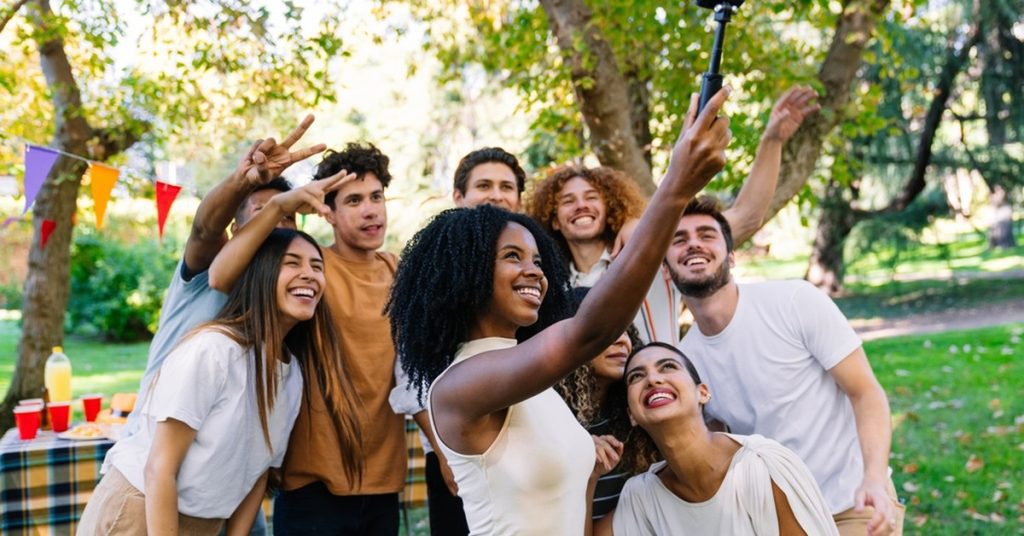 A diverse group of cheerful young adults poses for a selfie in a park near a picnic table filled with food and drinks.