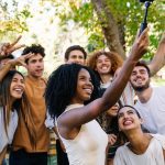 A diverse group of cheerful young adults poses for a selfie in a park near a picnic table filled with food and drinks.