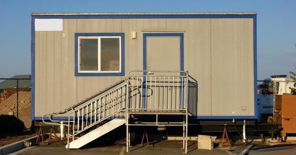 A tan portable office with blue trim has a metal staircase leading to a tan door. It has one window.