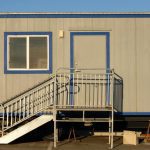 A tan portable office with blue trim has a metal staircase leading to a tan door. It has one window.
