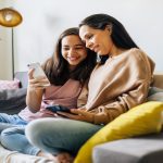 A mother and daughter sitting on a couch together, looking at the younger girl's phone. They are both smiling.