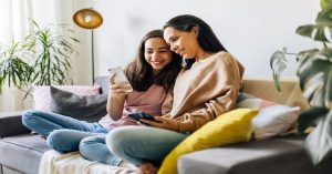 A mother and daughter sitting on a couch together, looking at the younger girl's phone. They are both smiling.
