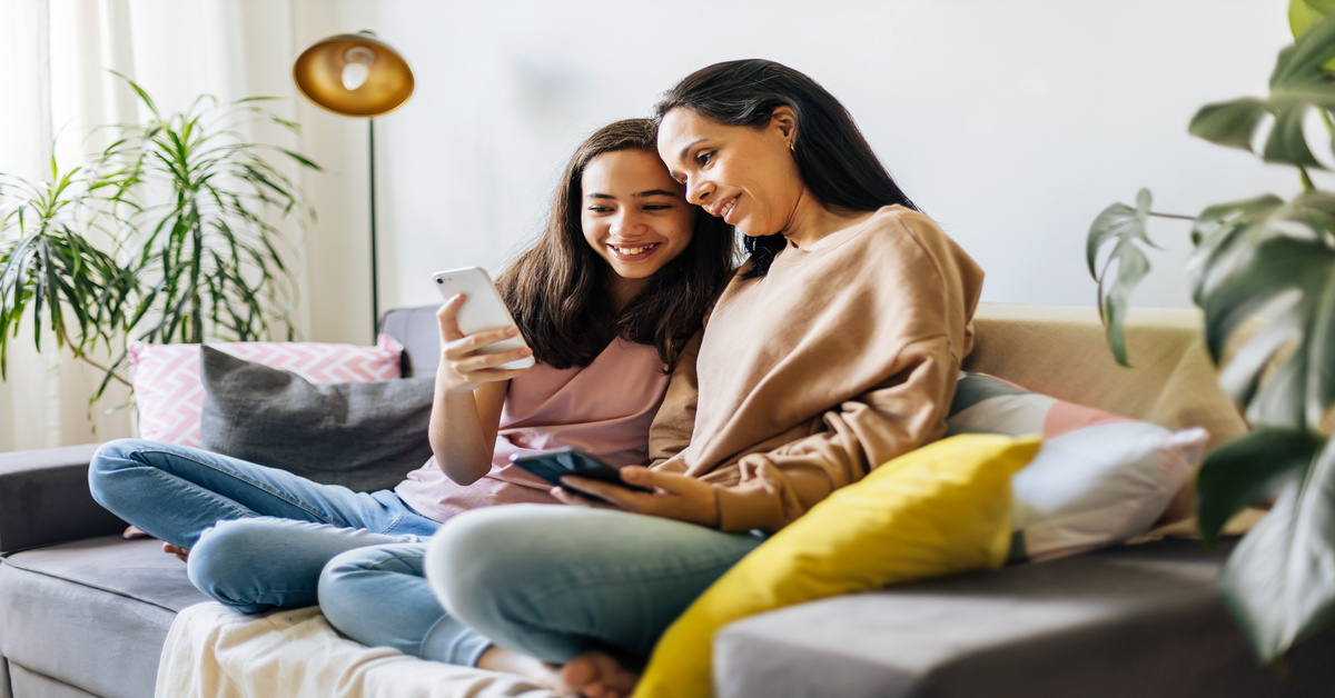 A mother and daughter sitting on a couch together, looking at the younger girl's phone. They are both smiling.