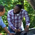 A father and his son smile together as they work underneath the hood of a car. Trees stand in the background.