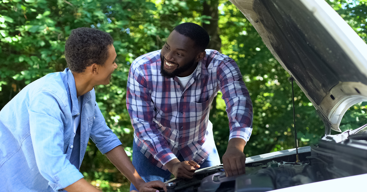 A father and his son smile together as they work underneath the hood of a car. Trees stand in the background.