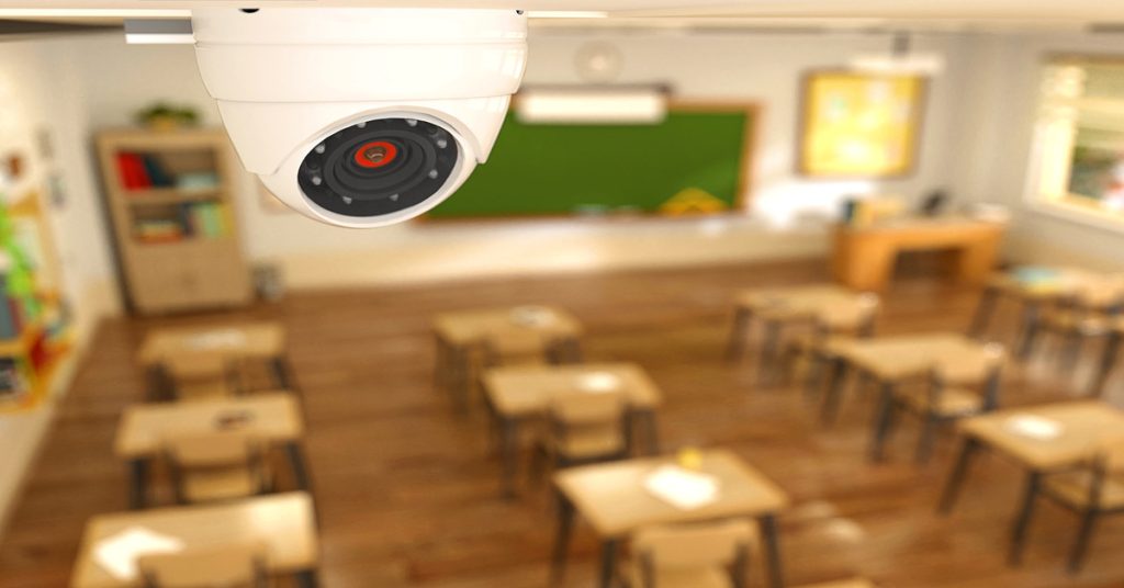 A white circular security camera mounted on a classroom wall above wooden desks and a green chalkboard.