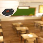 A white circular security camera mounted on a classroom wall above wooden desks and a green chalkboard.