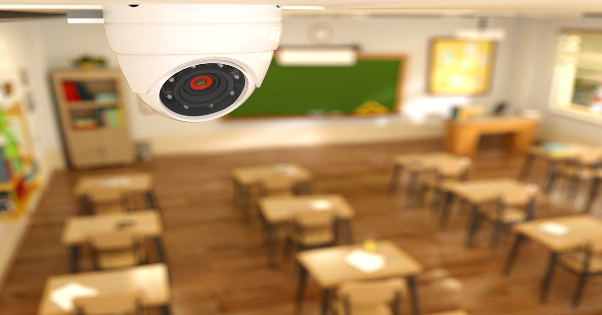 A white circular security camera mounted on a classroom wall above wooden desks and a green chalkboard.