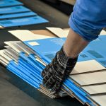 A person wearing a blue and gray shirt and black gloves is placing flat white and blue boxes on a black platform.