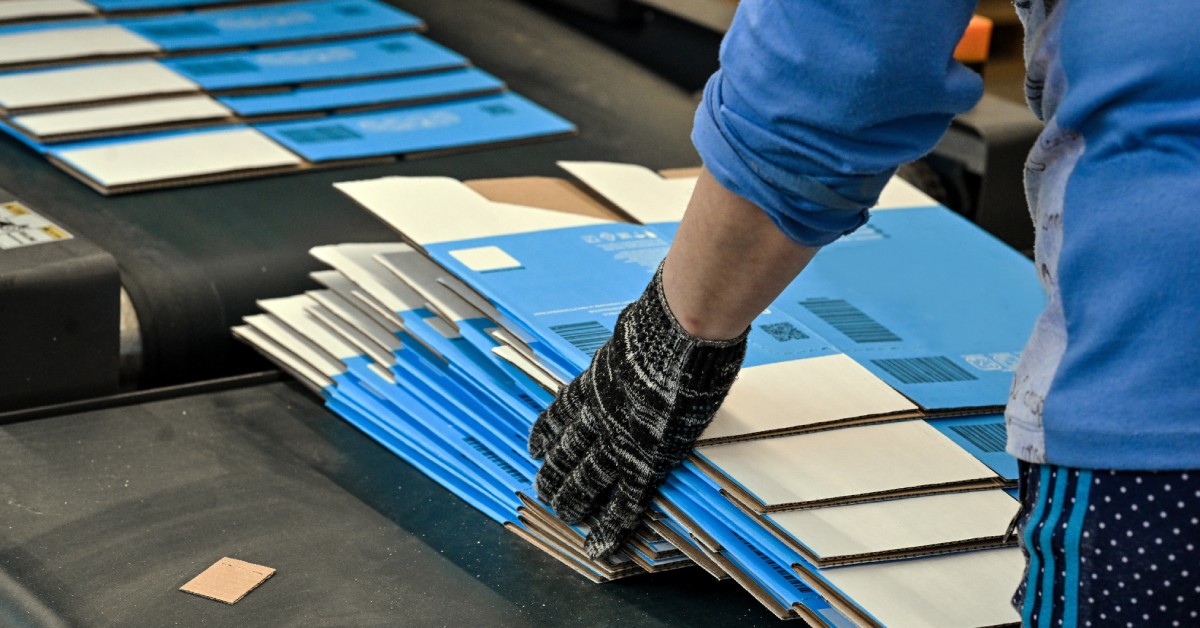 A person wearing a blue and gray shirt and black gloves is placing flat white and blue boxes on a black platform.