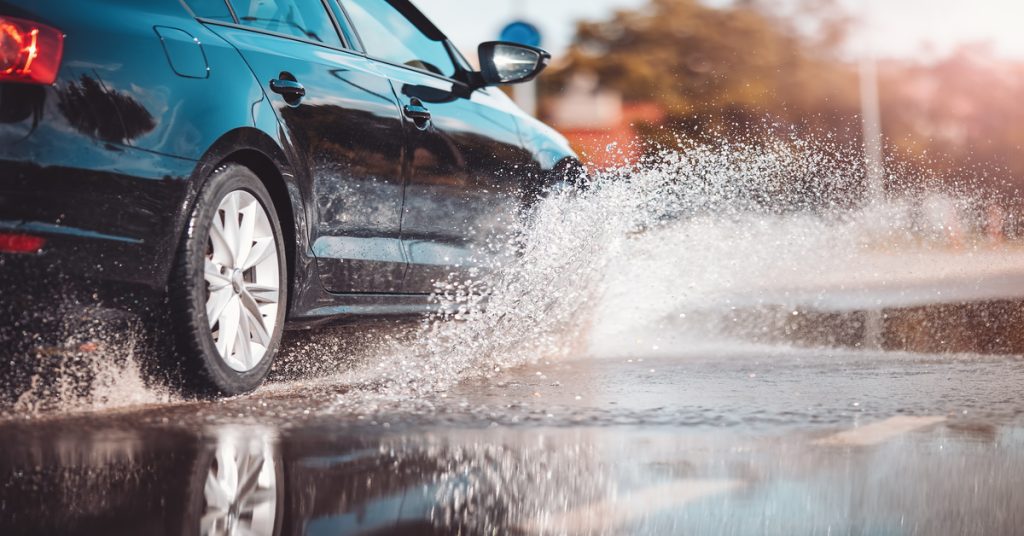 A black car driving through a puddle of water. Water is flying up into the air around the car's tire.