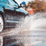 A black car driving through a puddle of water. Water is flying up into the air around the car's tire.