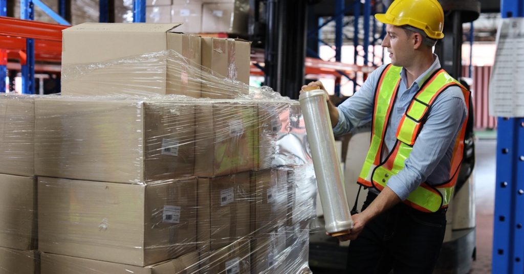 A man in a yellow hard hat and safety vest stretch wraps a pallet of cardboard boxes in a warehouse.